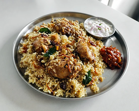 Chicken biriyani with kaima rice, fried onions, cashews and raisins on a steel plate with raita and pickle